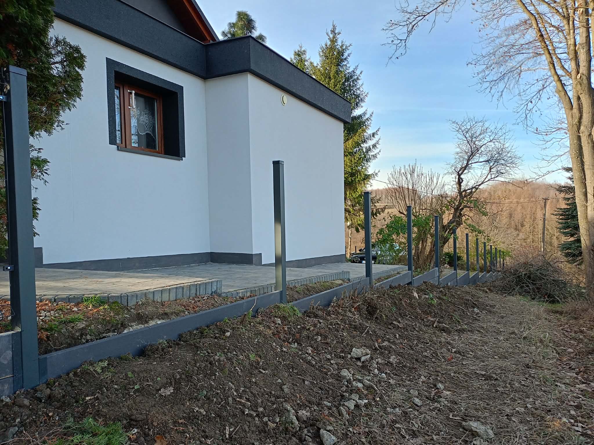 Wkopane słupki i podmurówka pod ogrodzenie panelowe na nierównym terenie, widok z boku, dom w tle, błękitne niebo.