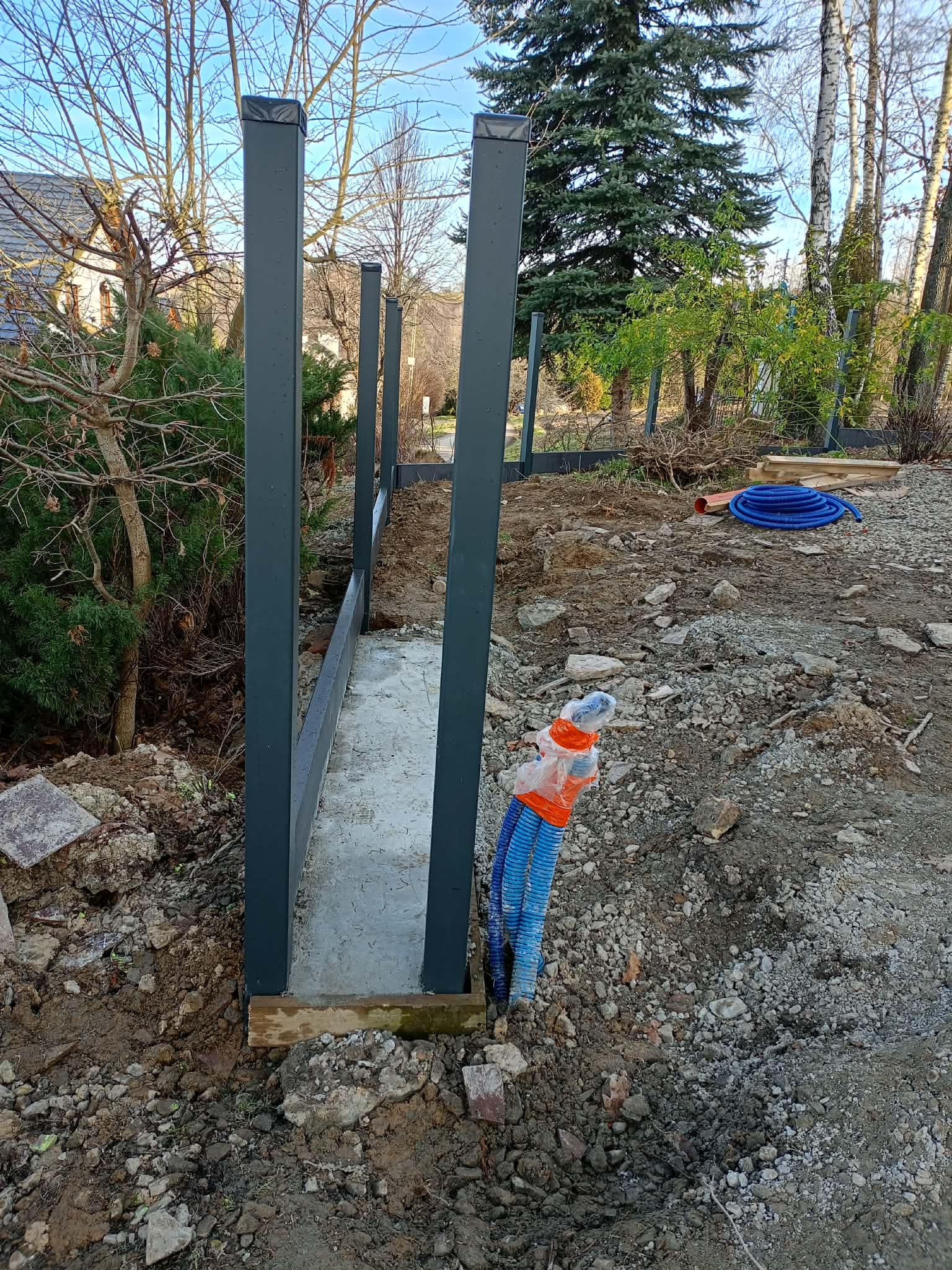 Wkopane słupki metalowe ogrodzenia na betonowej podmurówce, widoczne niebieskie rury w ziemi. Teren przygotowany pod montaż ogrodzenia.