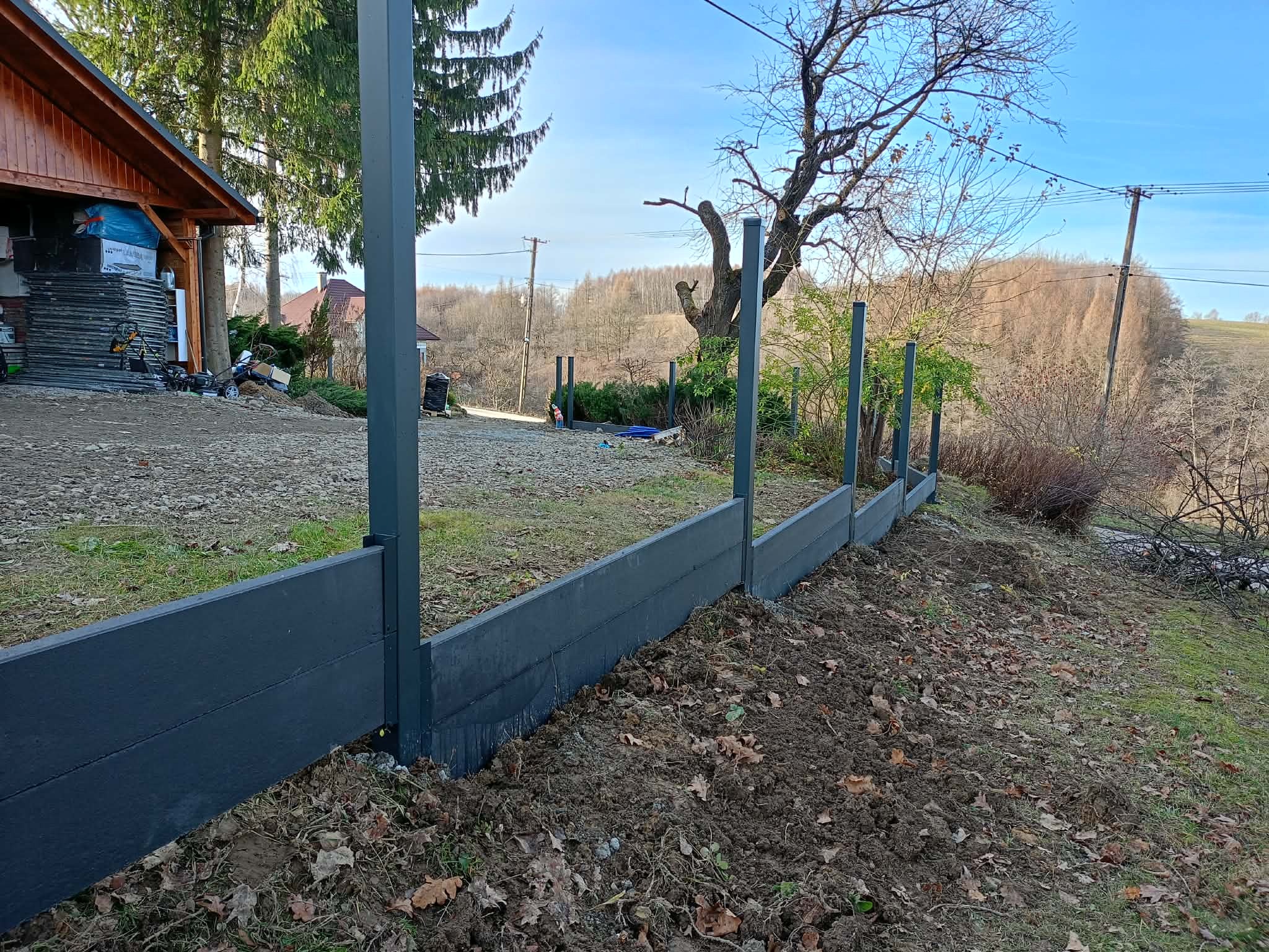 Częściowo ukończony, nowoczesny, grafitowy płot z paneli kompozytowych na nierównym terenie. Widoczne słupki i deski, w tle dom i drzewa.