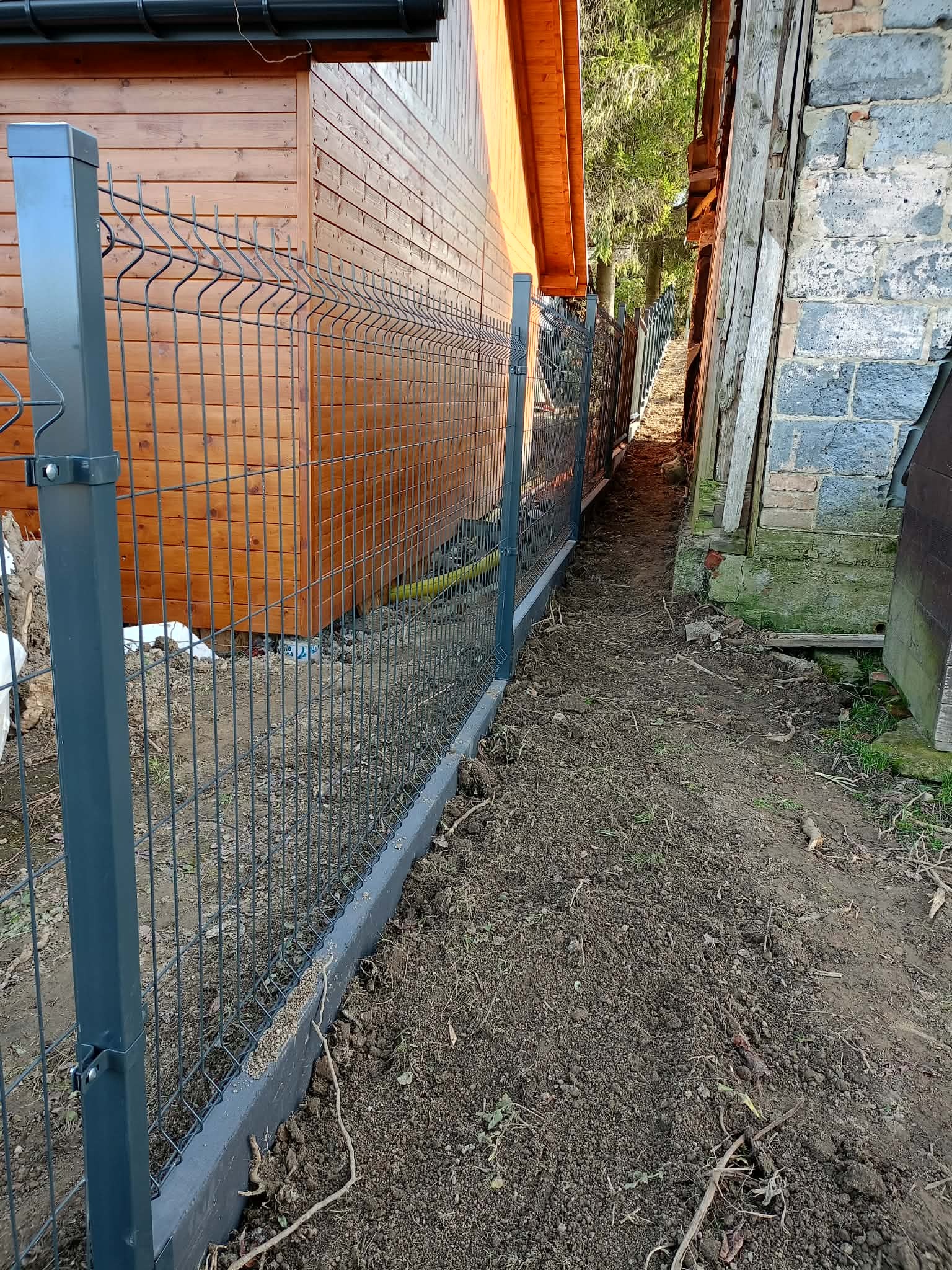 Nowoczesne ogrodzenie panelowe w kolorze grafitowym, zamontowane na betonowej podmurówce, oddzielające drewniany budynek od starego, murowanego budynku.