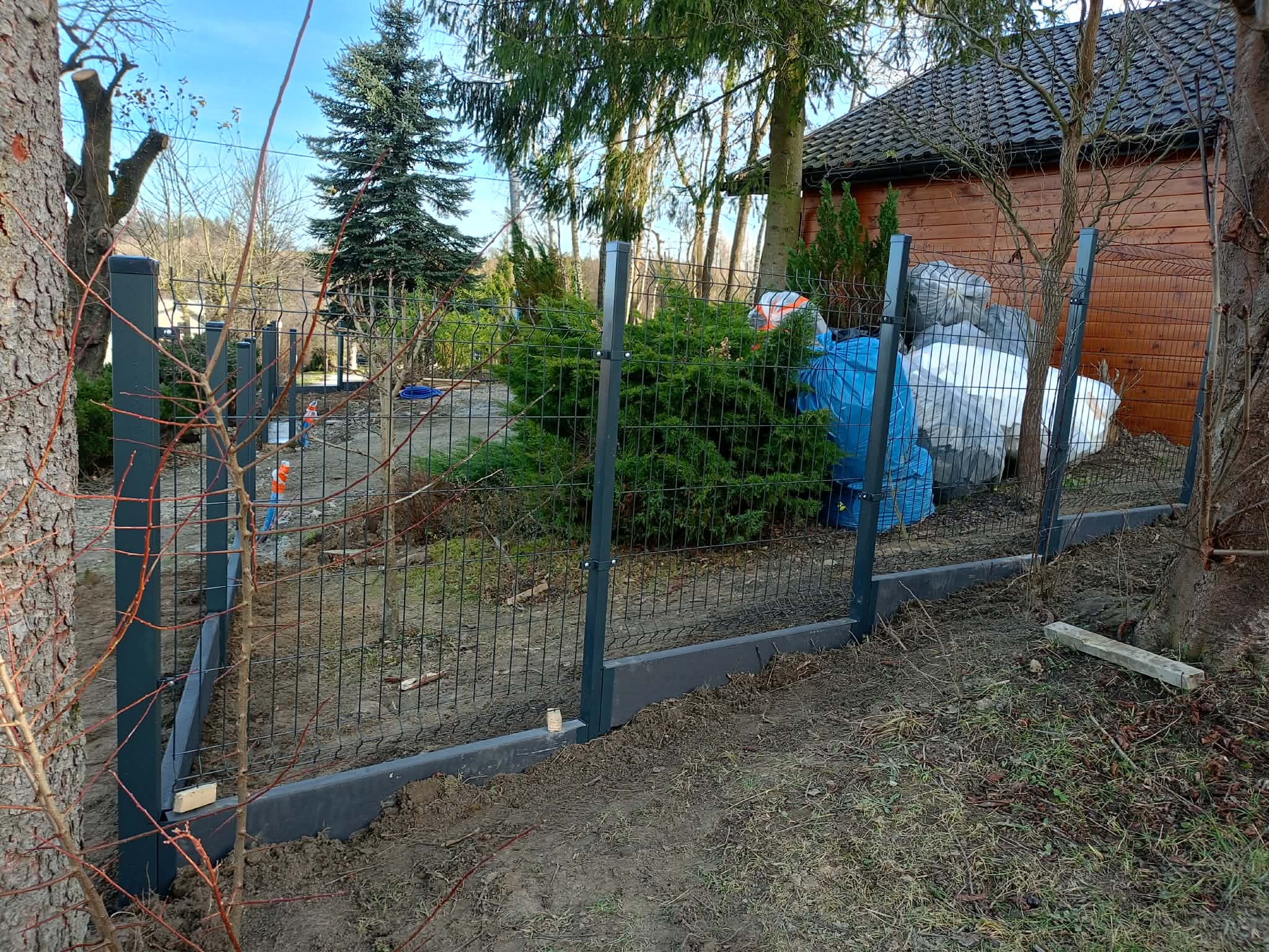 Montaż ogrodzenia panelowego na podmurówce betonowej. Widoczne słupki i siatka ogrodzeniowa w trakcie instalacji, z elementami mocującymi. Tło: ogród z drzewami i budynkiem.