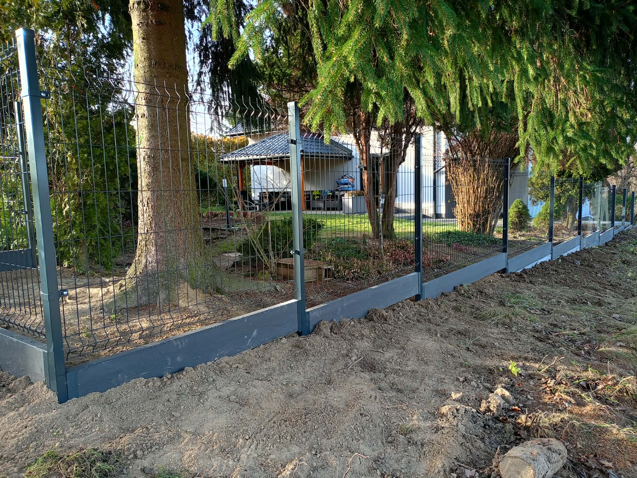 Nowoczesne ogrodzenie panelowe z podmurówką w Strzyżowie, widoczne słupki i siatka. Tło stanowią drzewa i zabudowania.