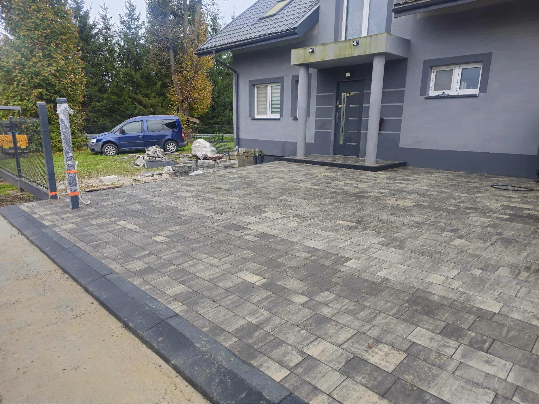 Ułożona kostka brukowa przed domem w odcieniach szarości, z widocznymi obrzeżami. W tle zaparkowany niebieski samochód i elementy przygotowane do montażu ogrodzenia.