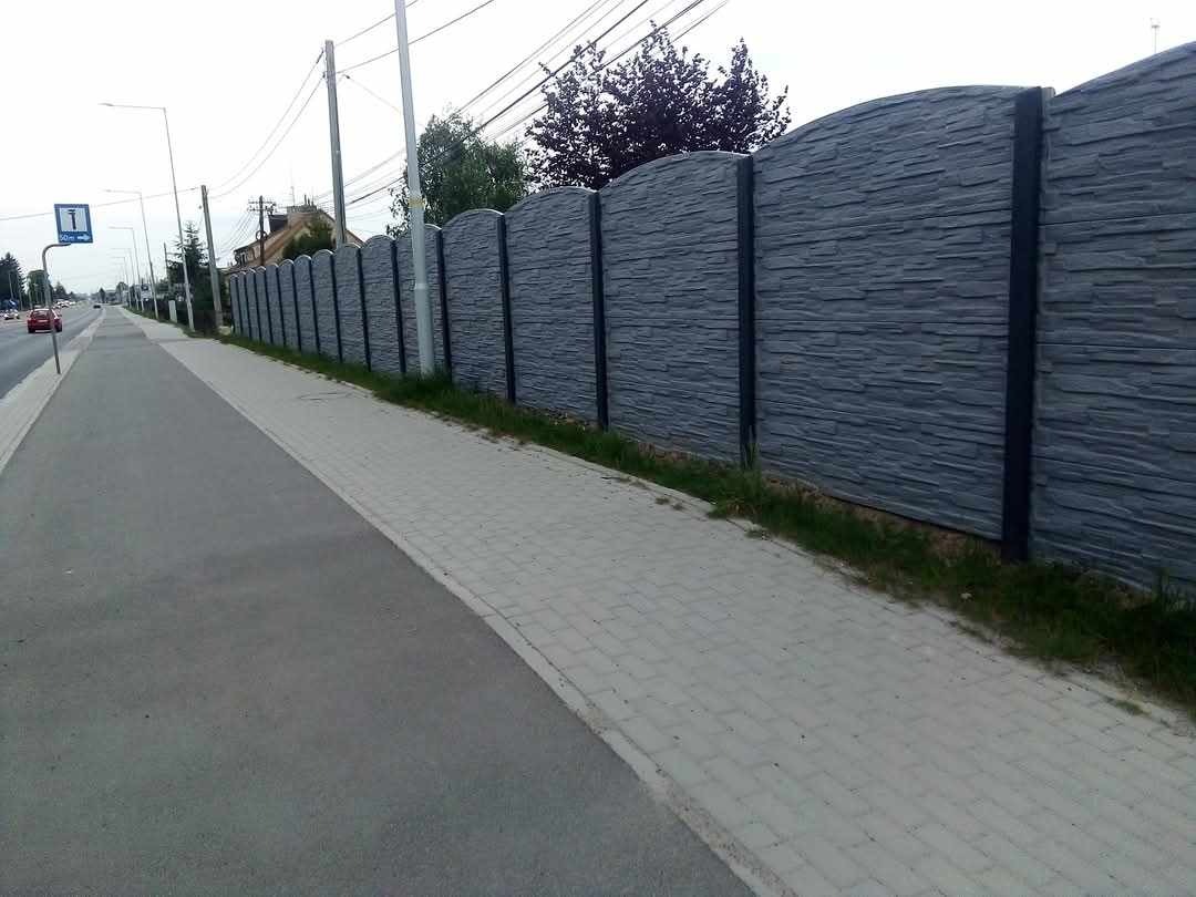 Długi, szary, łukowaty mur z fakturą kamienia, oddzielający chodnik od ulicy. Słupki w kolorze grafitowym. Widok z perspektywy przechodnia.