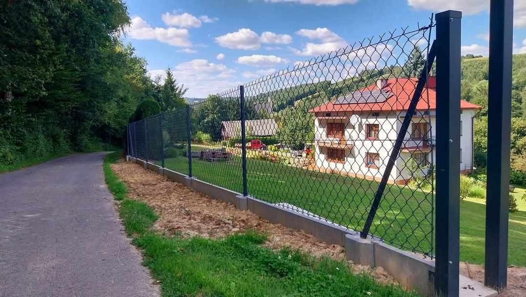 Nowoczesne ogrodzenie z siatki, czarne słupki, betonowa podmurówka, widok na dom z czerwonym dachem i panele słoneczne w tle, zielona trawa i drzewa.