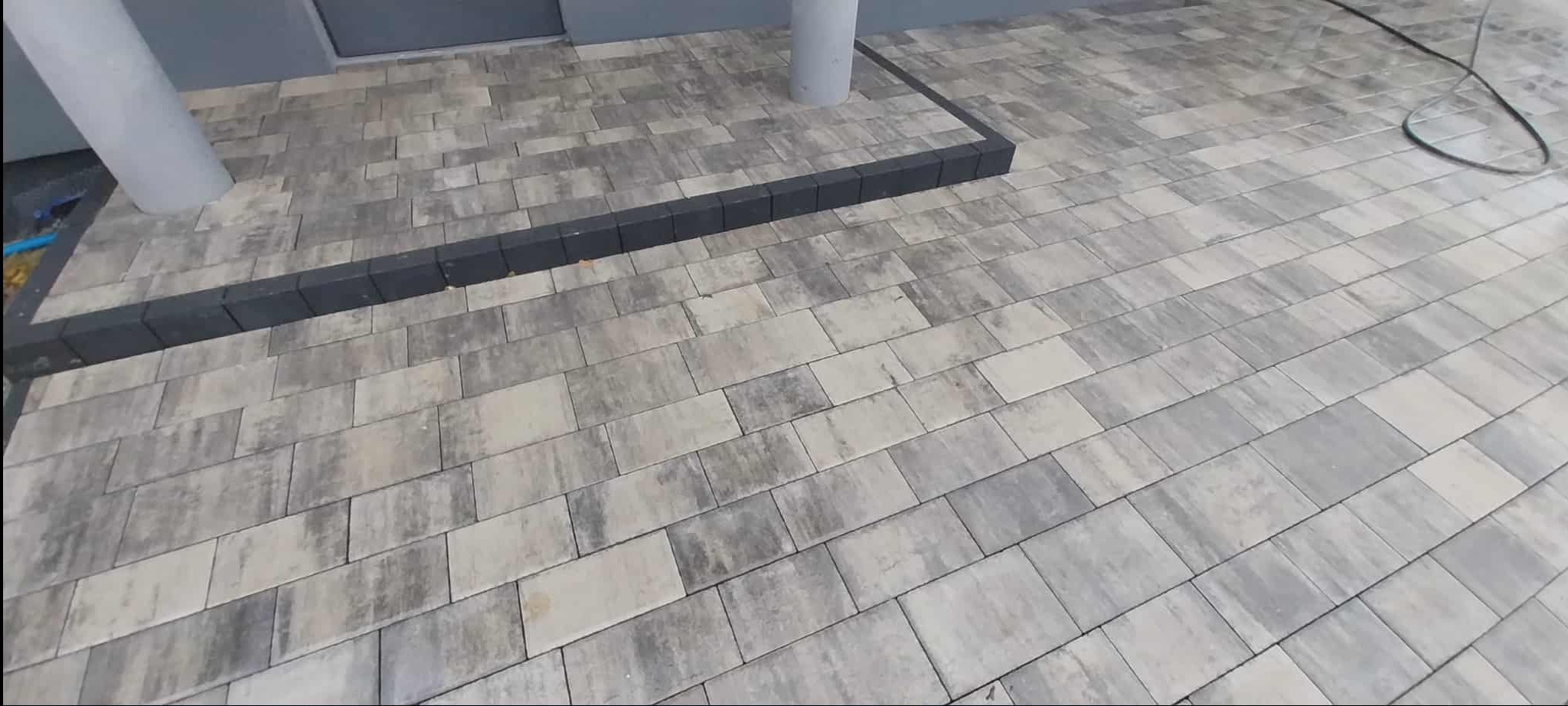 Ułożona kostka brukowa w odcieniach szarości, obramowana ciemniejszymi kostkami. Widoczne dwie kolumny budynku. Całość tworzy estetyczne wykończenie.