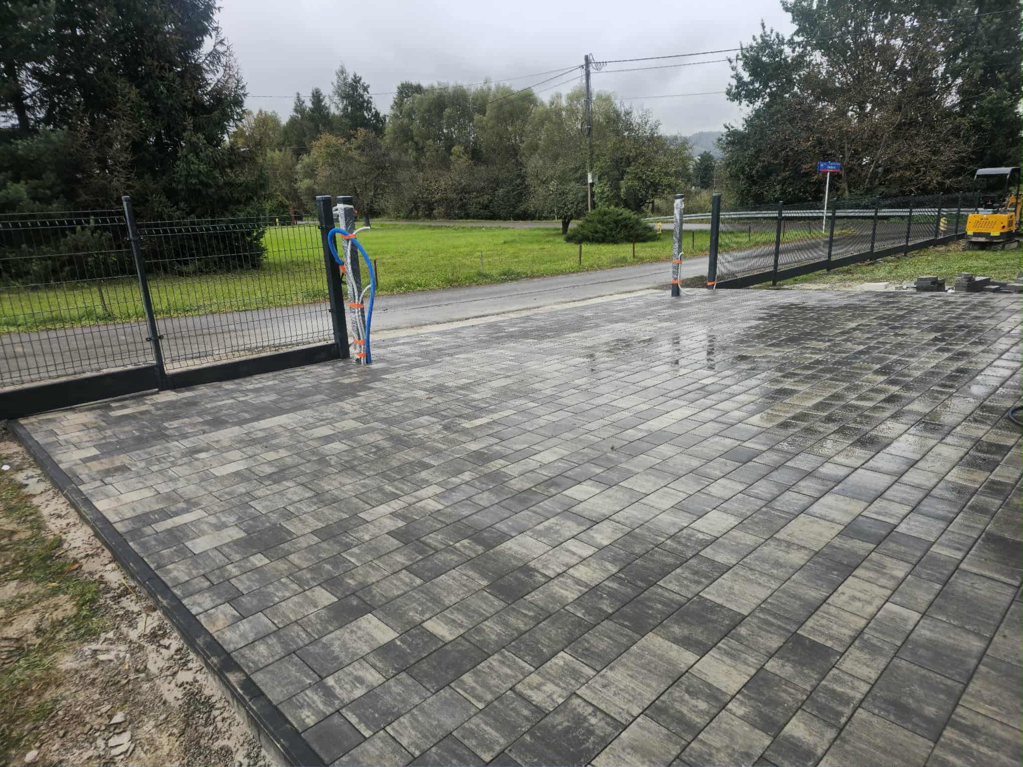 Ułożona kostka brukowa w szaro-beżowych odcieniach tworząca podjazd z bramą i ogrodzeniem panelowym. Widoczne słupki z instalacją elektryczną i fragment koparki.