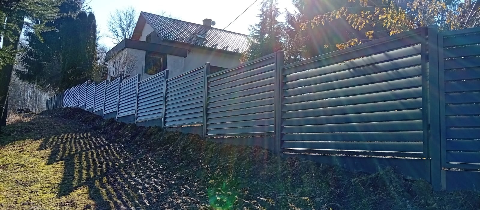 Nowoczesne, poziome ogrodzenie lamelowe w kolorze grafitowym, z widocznym domem w tle. Ujęcie z poziomu gruntu, podkreślające długość i estetykę ogrodzenia.