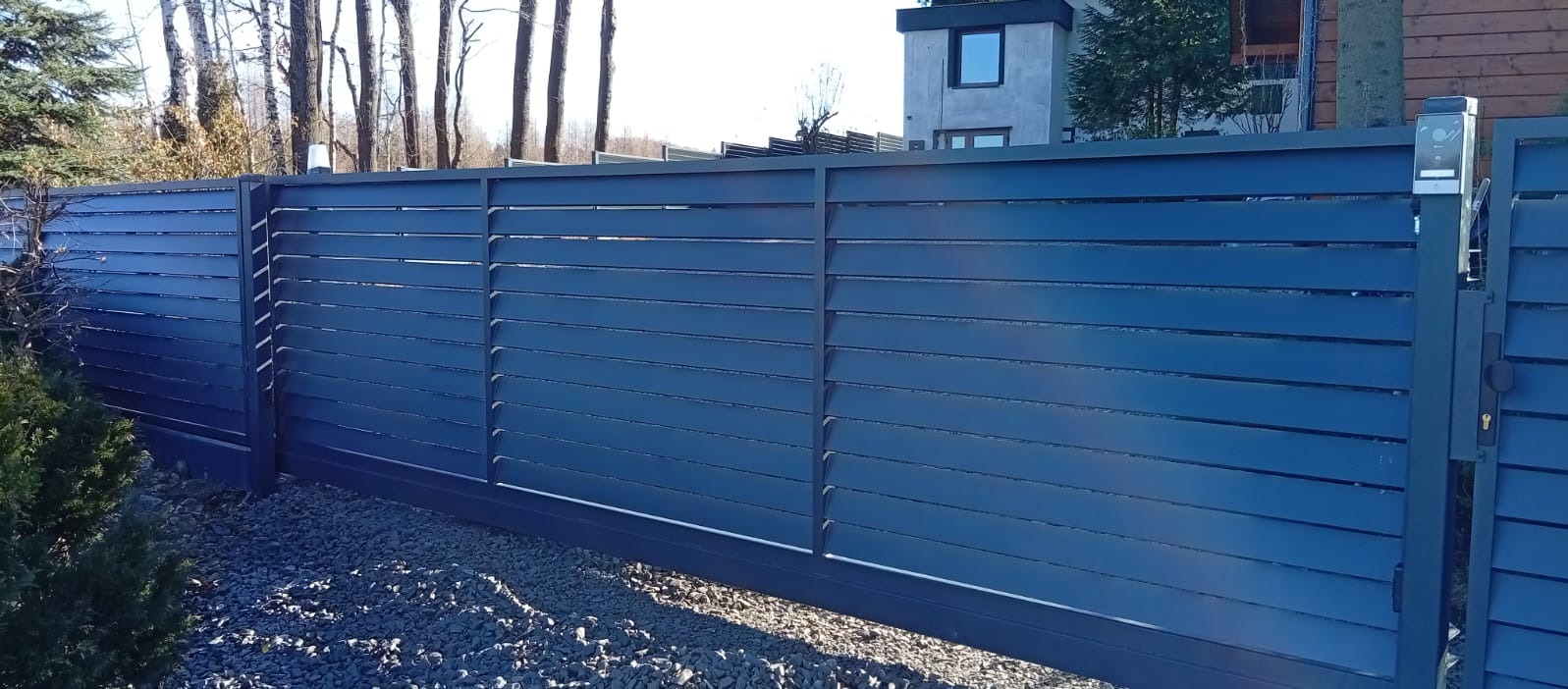 Nowoczesna, przesuwna brama metalowa w kolorze grafitowym z poziomymi lamelami, zamontowana na posesji z szarym żwirem. Widoczny fragment domu w tle.