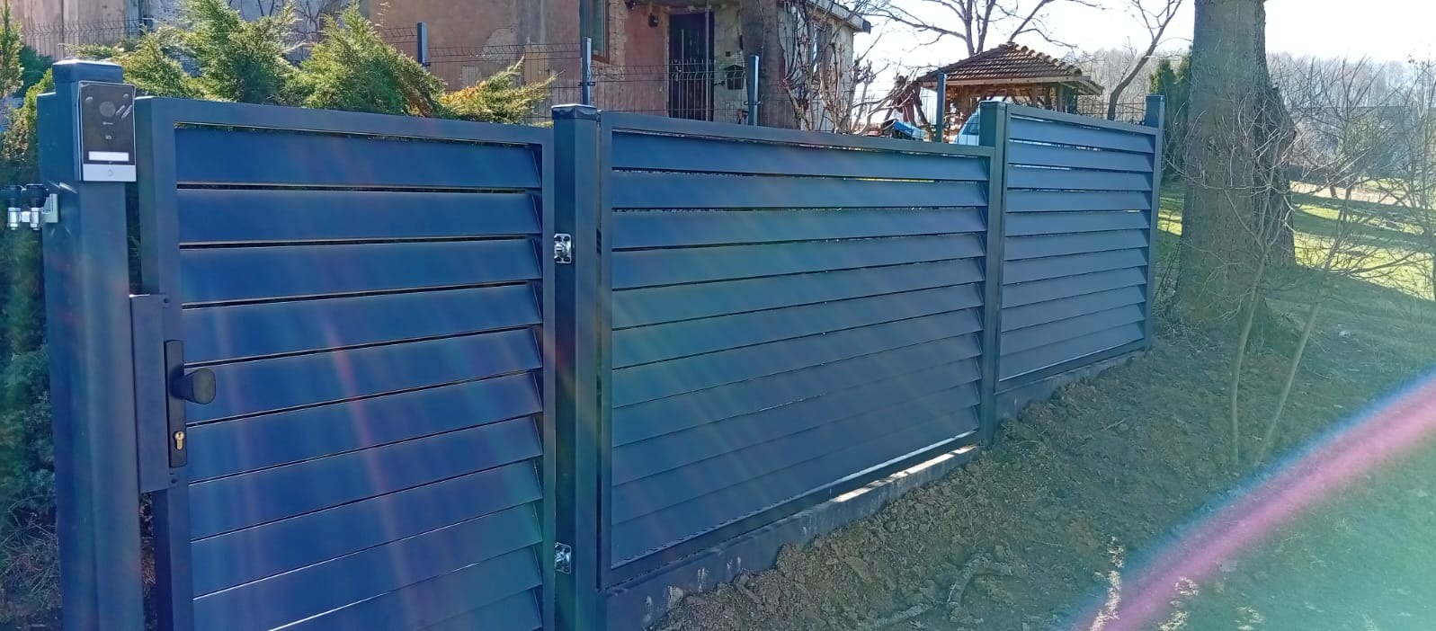 Nowoczesne, poziome ogrodzenie panelowe w kolorze grafitowym z furtką i wideodomofonem. Widoczne mocowania i detale wykończenia. Tło: dom i drzewa.