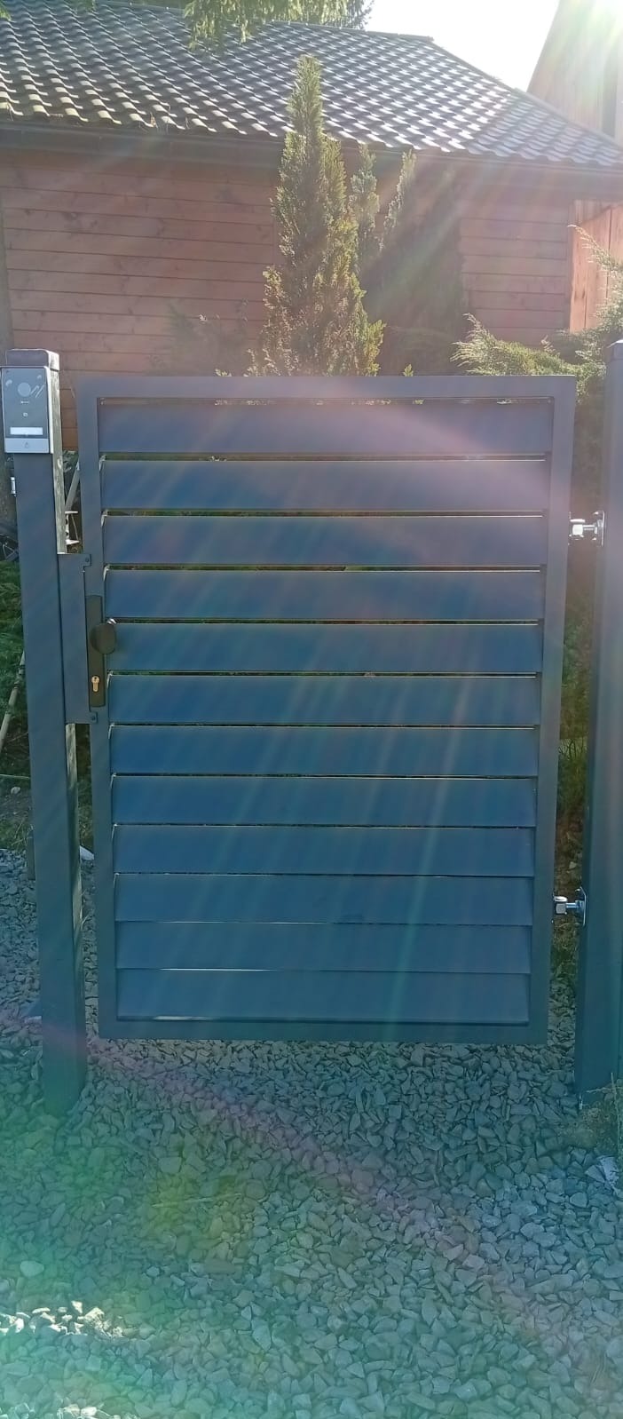 Nowoczesna, szara furtka lamelowa z domofonem, zamontowana na posesji z drewnianym domem w tle. Furtka osadzona na kamiennym podłożu.