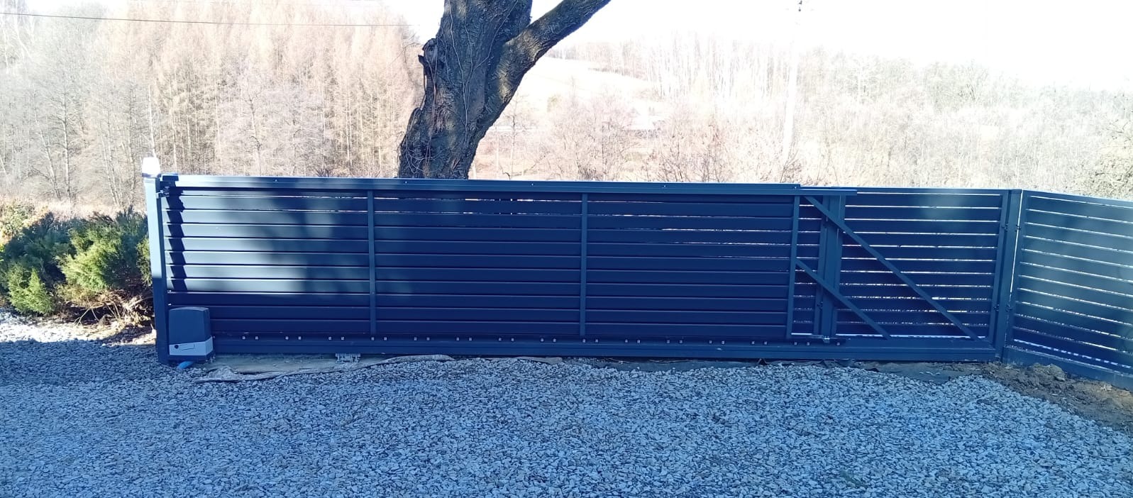 Nowoczesna, przesuwna brama posesyjna z poziomych paneli w kolorze grafitowym, z widocznym napędem, na tle drzewa i żwirowego podłoża w słoneczny dzień.