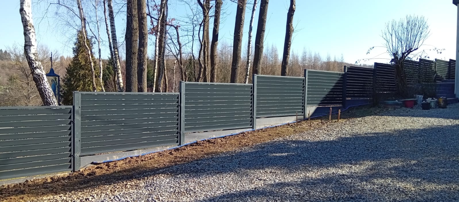 Nowoczesne, poziome ogrodzenie panelowe w kolorze grafitowym, zamontowane na betonowej podmurówce. Widoczne drzewa w tle i żwirowe podłoże.