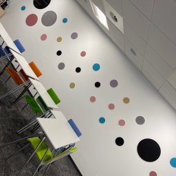 Białe biuro z kolorowymi krzesłami i okrągłymi dekoracjami na ścianie. Widok na sufit kasetonowy i stoliki. Nietypowy kąt ujęcia podkreśla nowoczesny design.