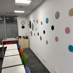 Biała ściana biura z dekoracyjnymi, kolorowymi kółkami. W tle widoczne stoły, krzesła i ekspres do kawy. Nowoczesny design wnętrza.