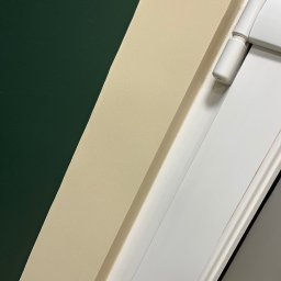 Fragment pomalowanej ściany na ciemnozielono, z beżową listwą przypodłogową i białymi drzwiami z zawiasem. Detal wykończenia wnętrza po malowaniu.