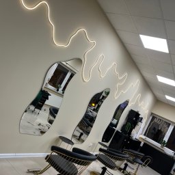 Nowoczesne wnętrze salonu fryzjerskiego z asymetrycznymi lustrami, oświetleniem LED i czarno-złotymi krzesłami. Widok na stanowiska pracy i okno.