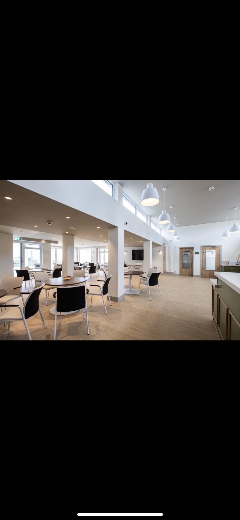 Jasne, przestronne wnętrze kawiarni/restauracji z drewnianą podłogą, białymi ścianami i kolumnami. Widoczne stoliki z krzesłami, lampy wiszące i fragment baru.