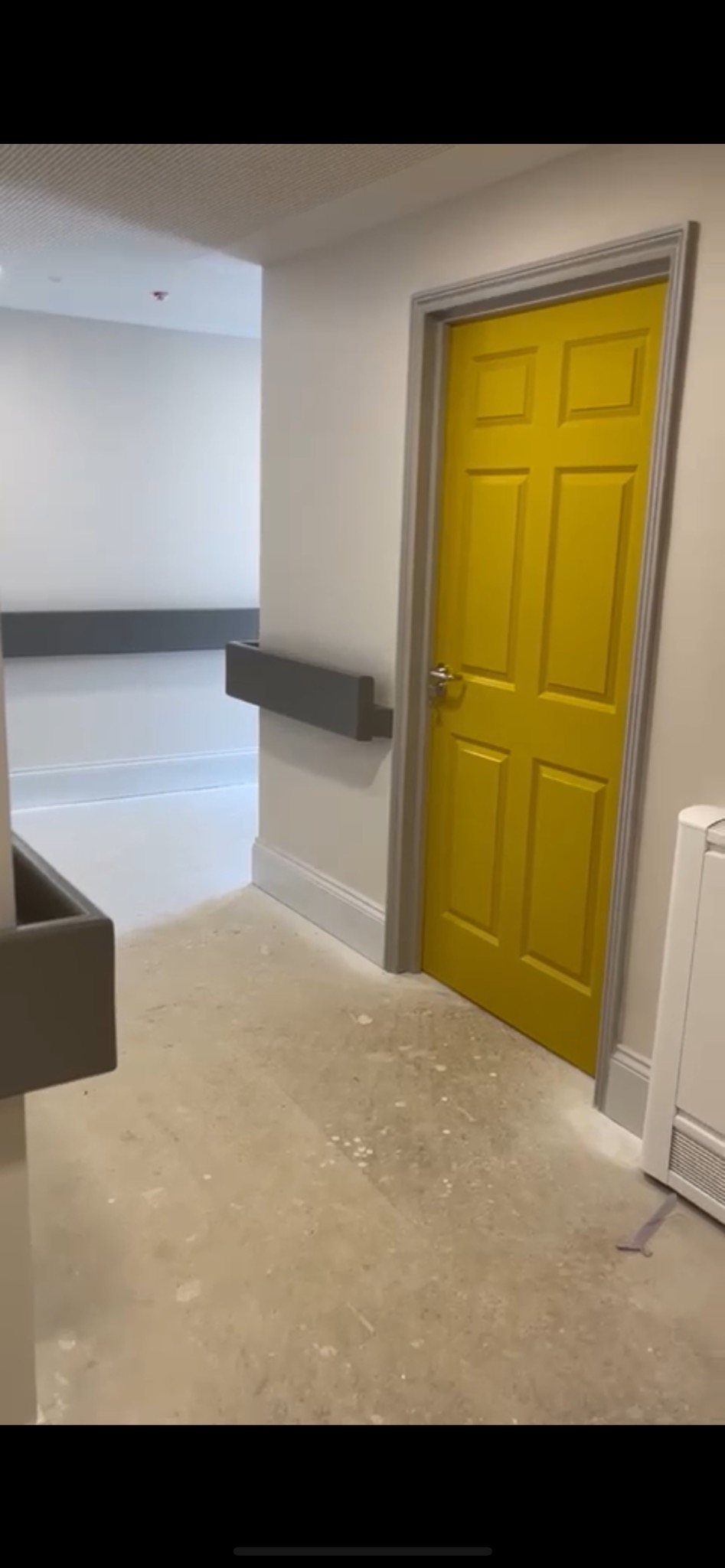 Żółte drzwi w szarej ramie w korytarzu z szarymi poręczami. Podłoga z plamami farby. Świeżo pomalowane ściany na biało.