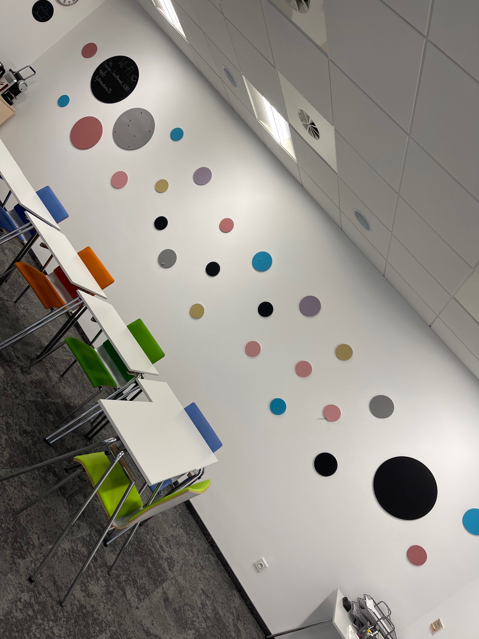 Białe biuro z kolorowymi krzesłami i okrągłymi dekoracjami na ścianie. Widok na sufit kasetonowy i stoliki. Nietypowy kąt ujęcia podkreśla nowoczesny design.