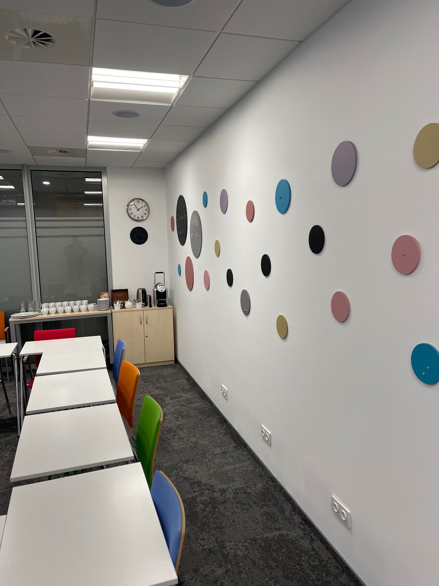 Biała ściana biura z dekoracyjnymi, kolorowymi kółkami. W tle widoczne stoły, krzesła i ekspres do kawy. Nowoczesny design wnętrza.
