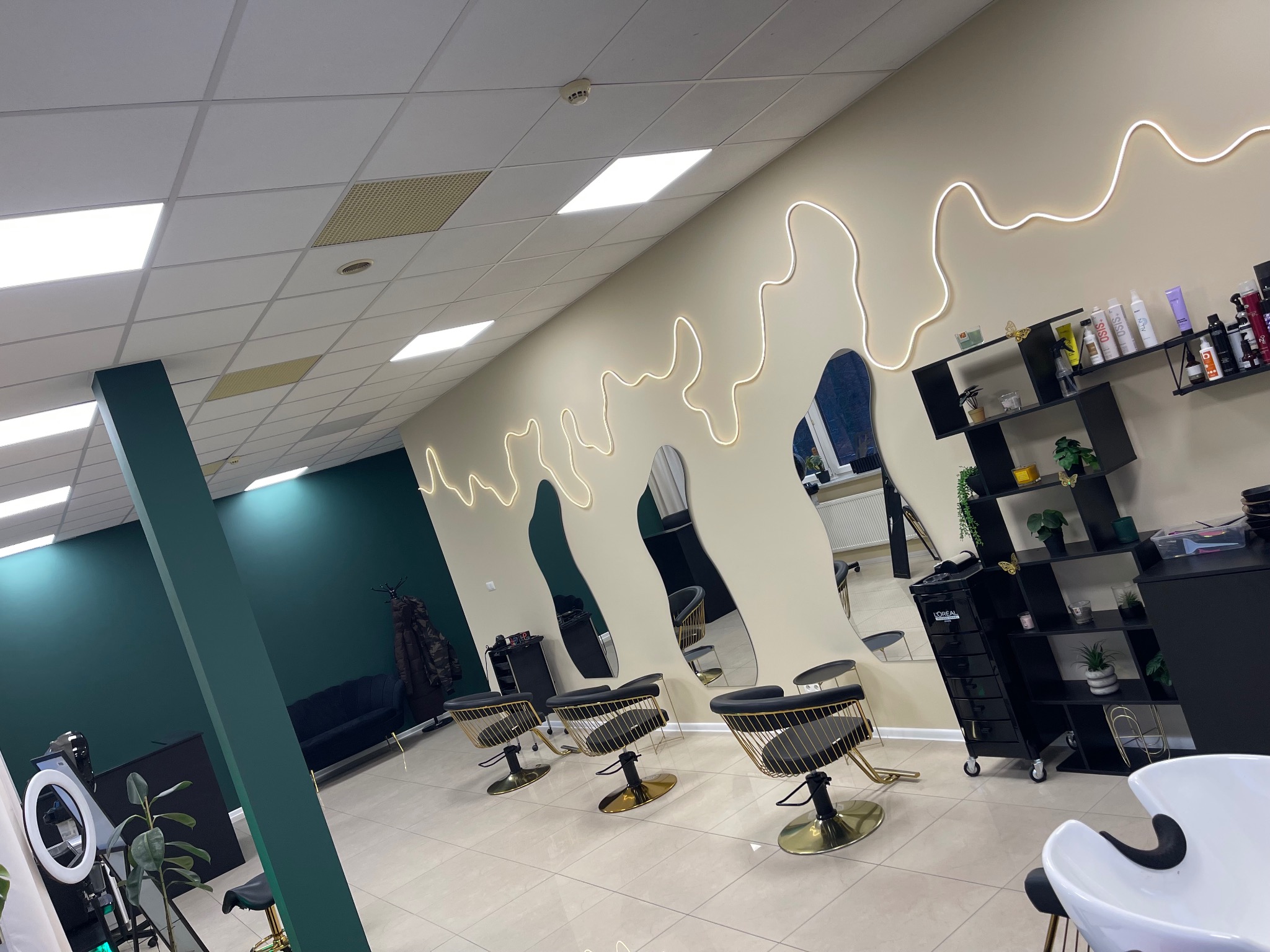 Nowoczesne wnętrze salonu fryzjerskiego z trzema stanowiskami, lustrami o nieregularnym kształcie i dekoracyjnym oświetleniem LED. Dominują kolory: biel, czerń i złoto.