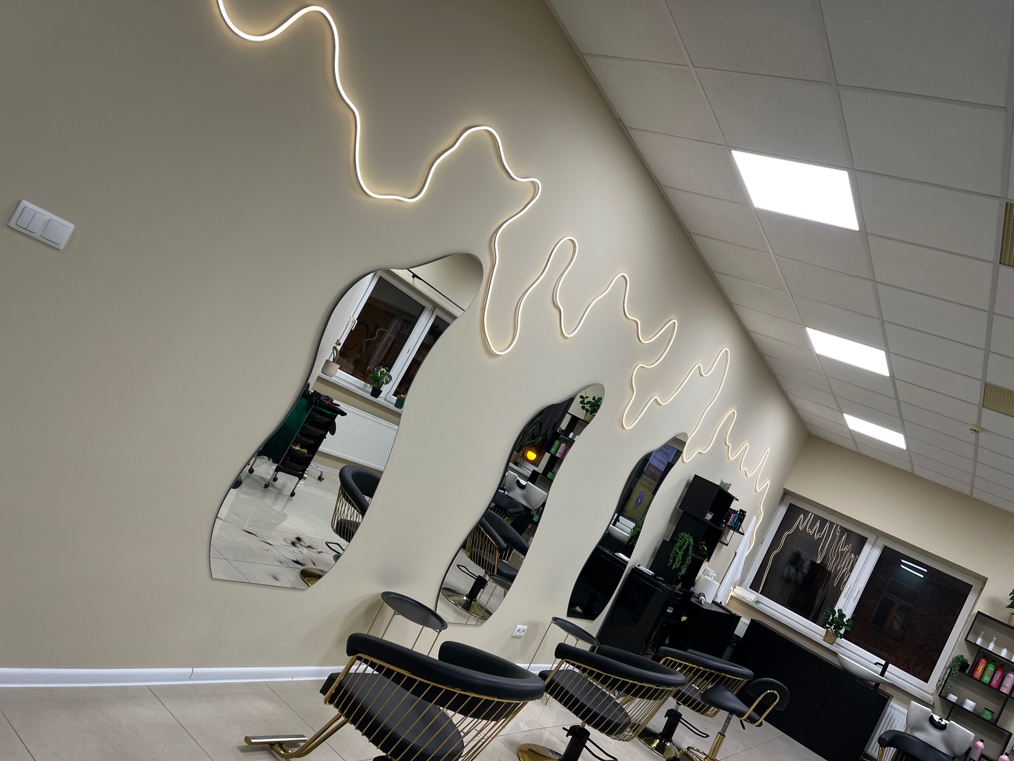 Nowoczesne wnętrze salonu fryzjerskiego z asymetrycznymi lustrami, oświetleniem LED i czarno-złotymi krzesłami. Widok na stanowiska pracy i okno.