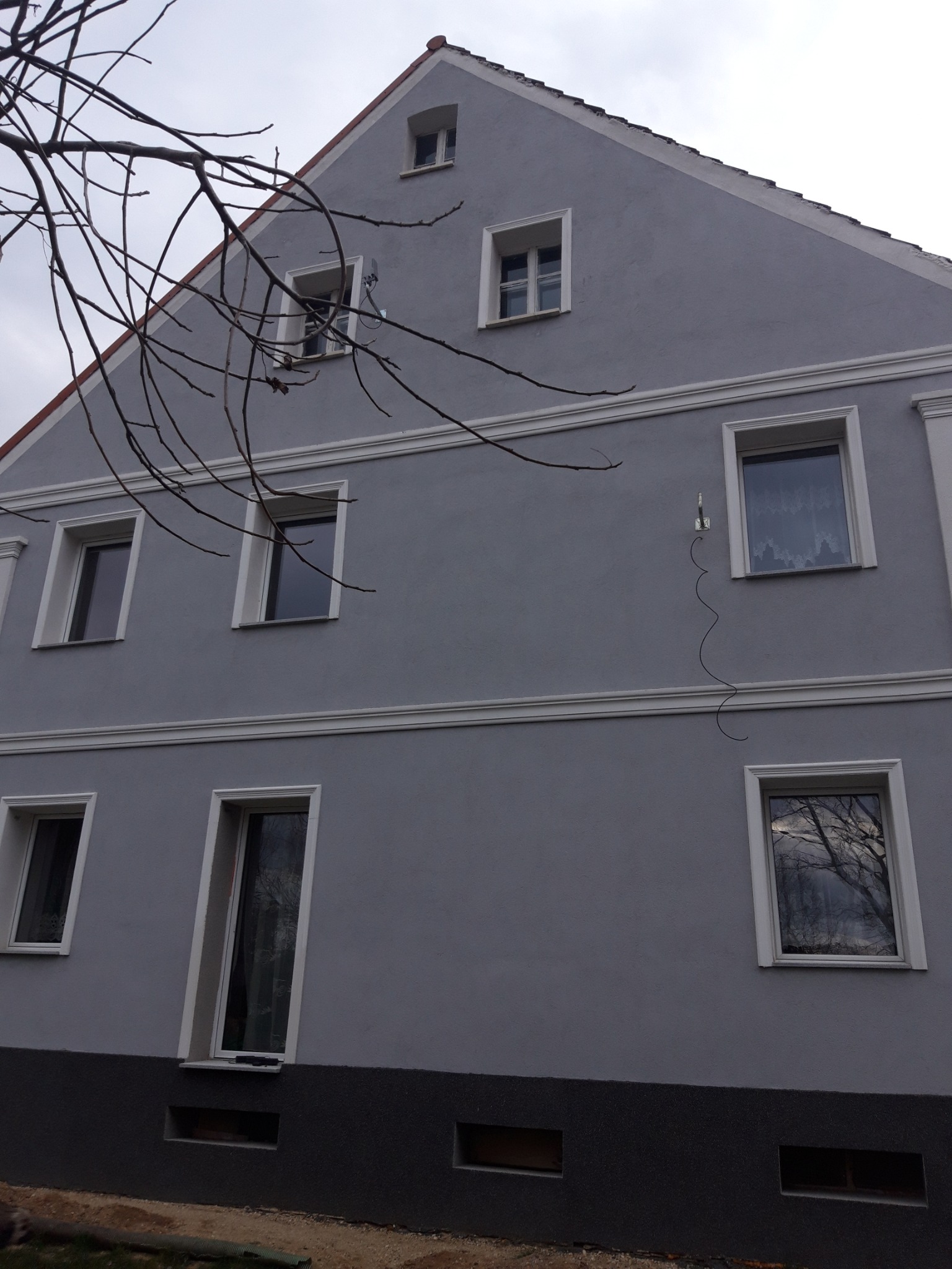 Szary budynek z białymi ramami okiennymi i dekoracyjnymi listwami. Widoczne nagie gałęzie drzewa w lewym górnym rogu. Nierównomierne oświetlenie.