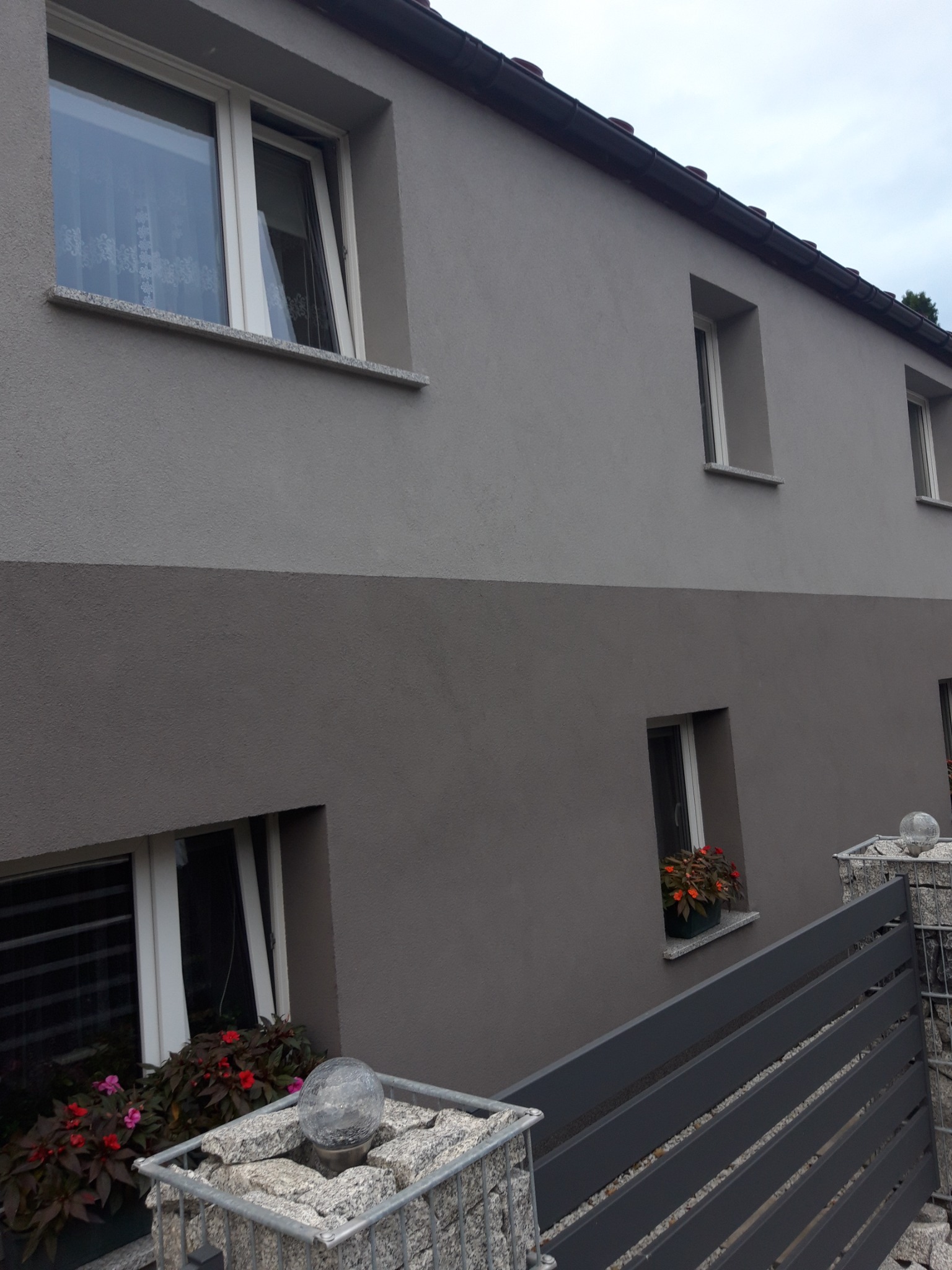 Dwukolorowa elewacja domu z oknami, kwiatami w donicach i ogrodzeniem z gabionów. Ujęcie z dołu, podkreślające detale wykończenia.