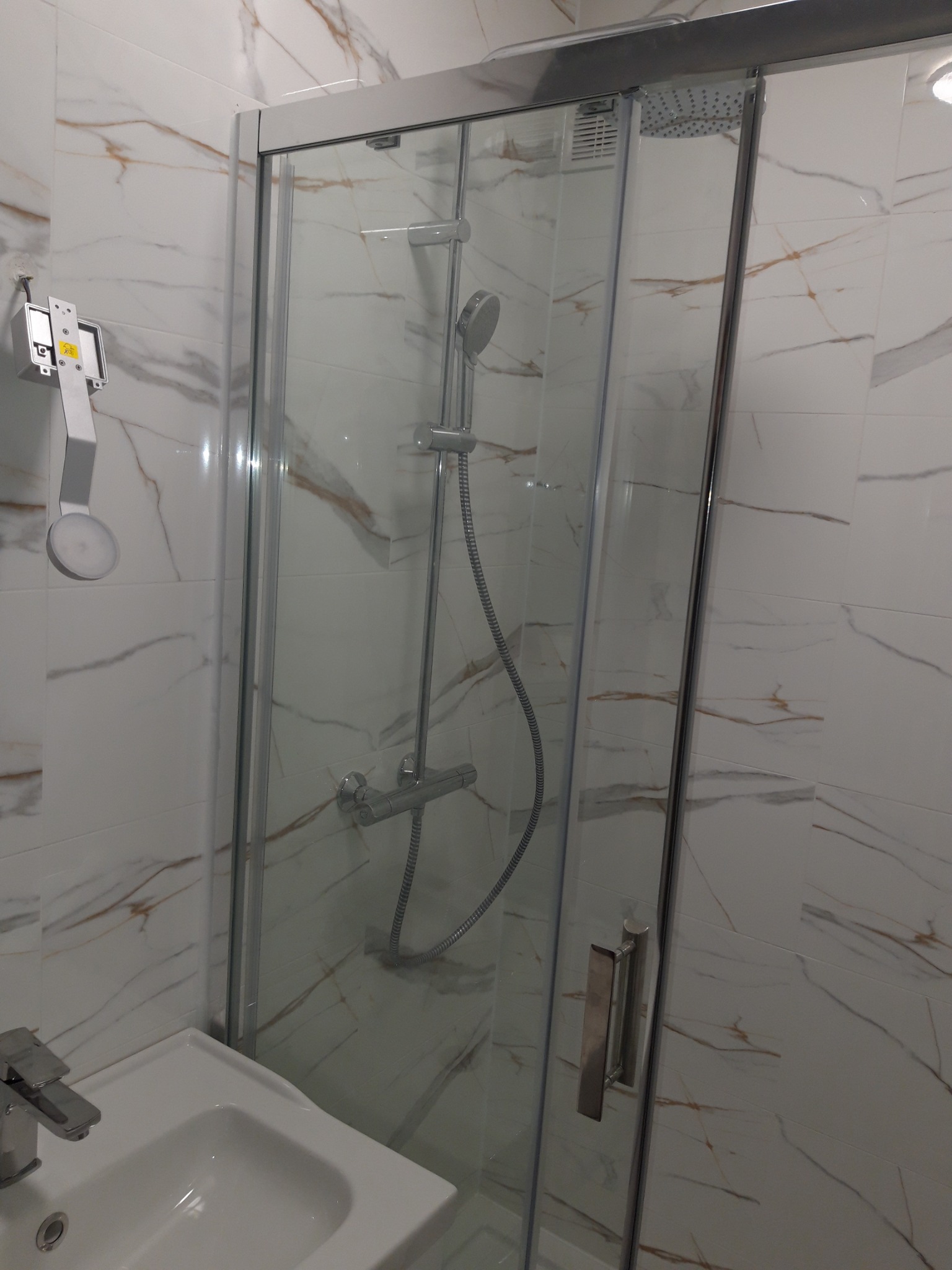 Nowoczesna kabina prysznicowa z przesuwnymi drzwiami, biała umywalka i marmurowe płytki w łazience. Widoczny chromowany zestaw natryskowy.