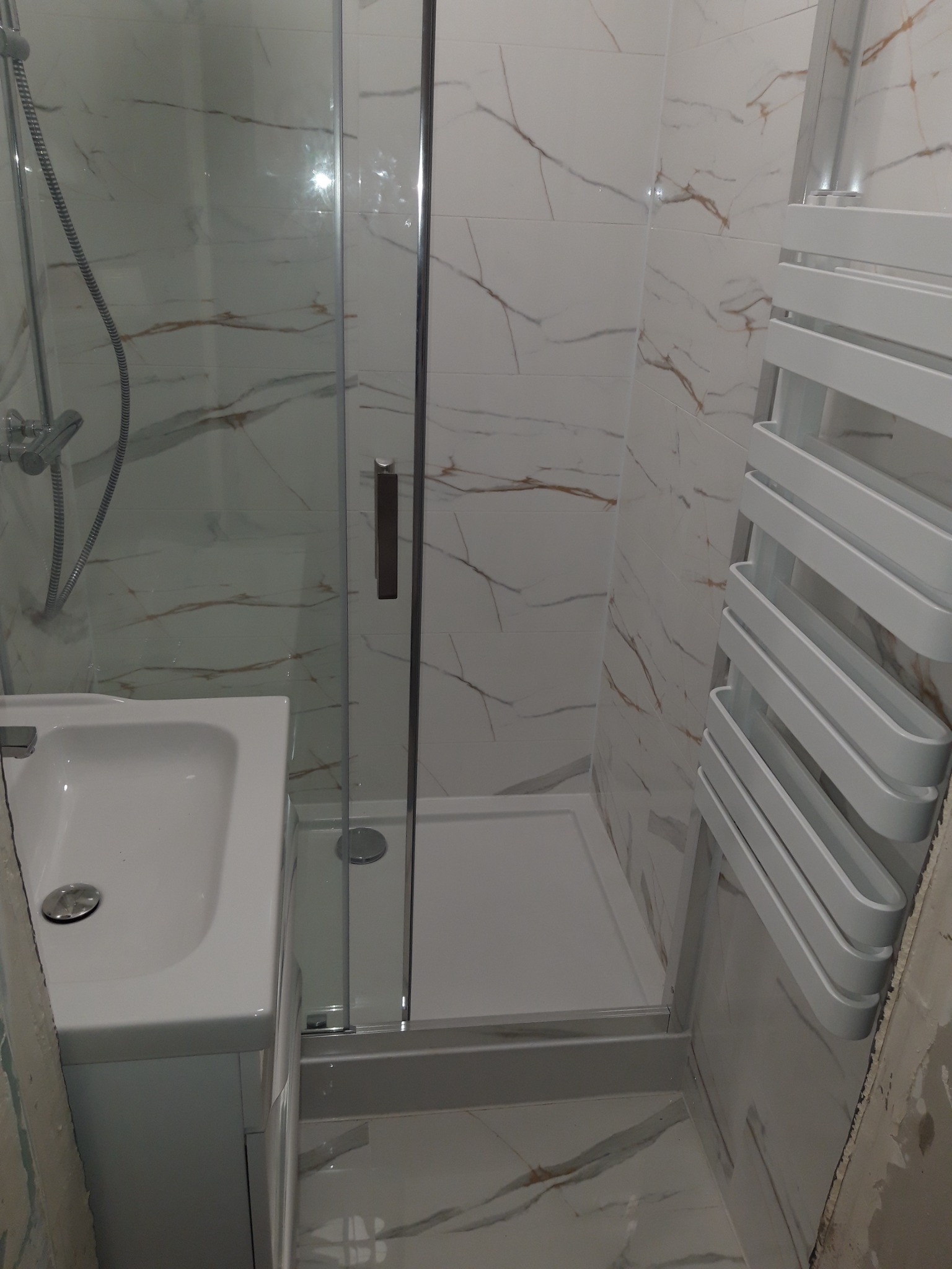 Wykończona łazienka z kabiną prysznicową, umywalką i grzejnikiem drabinkowym. Białe płytki z marmurkowym wzorem na ścianach i podłodze. Nowoczesny design.