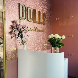 C&T Cichal - Eleganckie wnętrze: różowa mozaika heksagonalna na ścianie, złote litery 'DOLLS', biała lada recepcyjna z kwiatami, szafa w odcieniach różu.