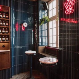 C&T Cichal - Stylowe wnętrze baru z ciemnymi, pionowo ułożonymi płytkami, drewnianą podłogą w jodełkę i neonowym napisem. Widoczne stoliki barowe i okno z rośliną.