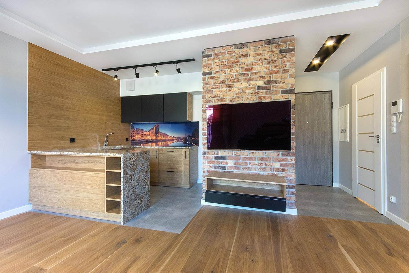 Nowoczesna aranżacja wnętrza: drewniana podłoga, ceglana ściana z TV, zabudowa kuchenna z blatem z granitu i fototapetą z widokiem na miasto. Oświetlenie punktowe.