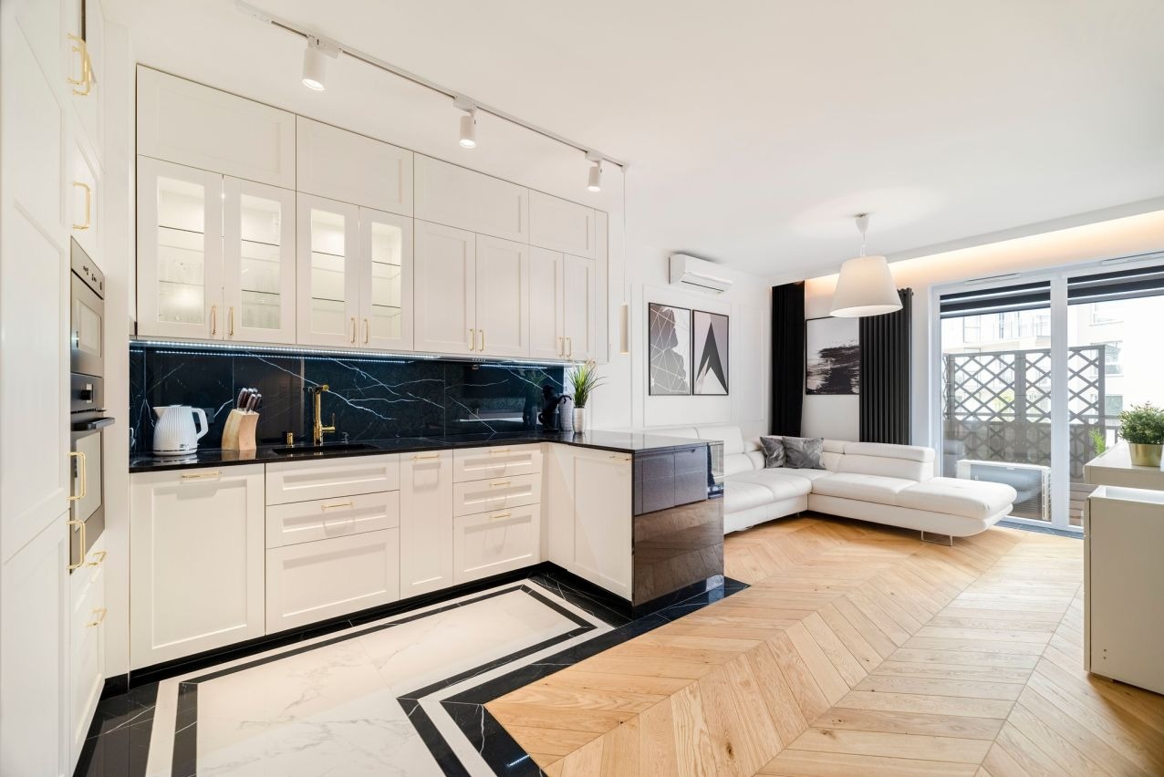 Elegancka kuchnia z białymi szafkami, czarnym blatem i marmurowym backsplashiem, połączona z salonem z parkietem w jodełkę i białą sofą narożną.