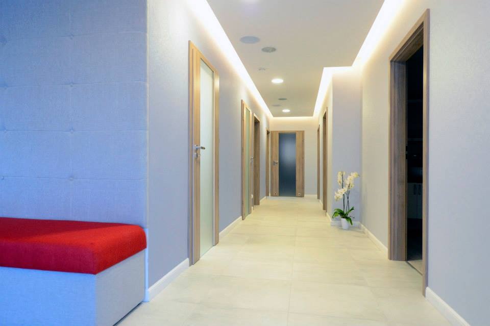 Korytarz biurowy po remoncie: jasne ściany, podświetlany sufit, drzwi z drewnianymi ościeżnicami, biała podłoga z płytek, czerwona ławka i kwiaty.