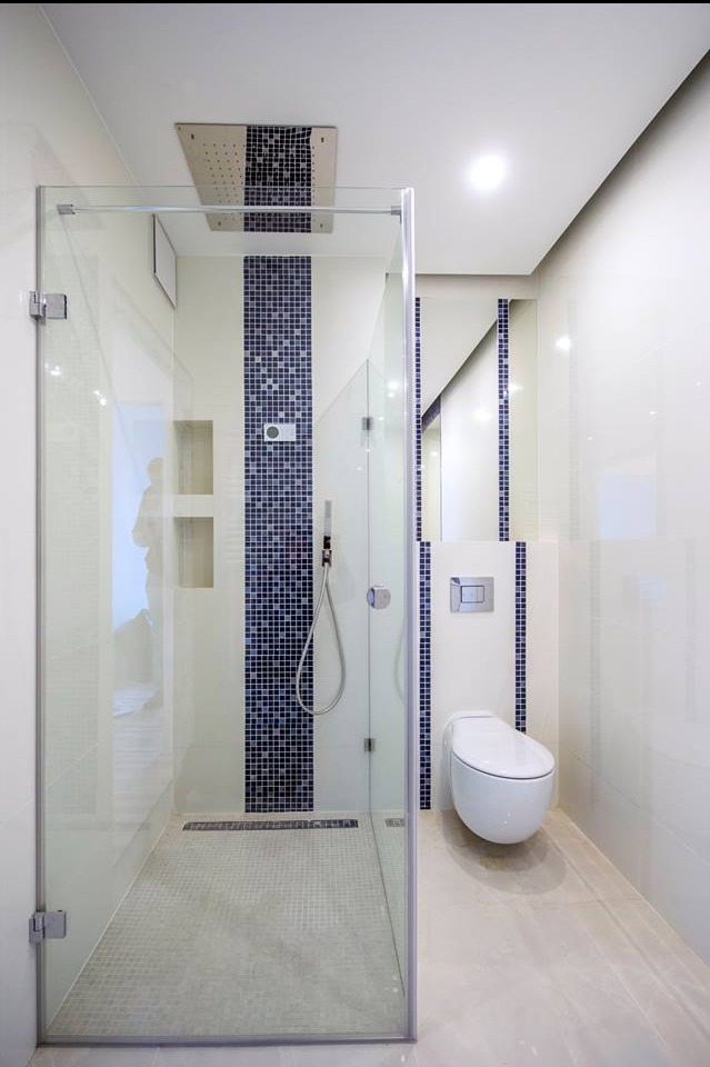 Nowoczesna łazienka z kabiną prysznicową typu walk-in, wykończona białymi płytkami i mozaiką w odcieniach granatu. Widoczna toaleta podwieszana i deszczownica.