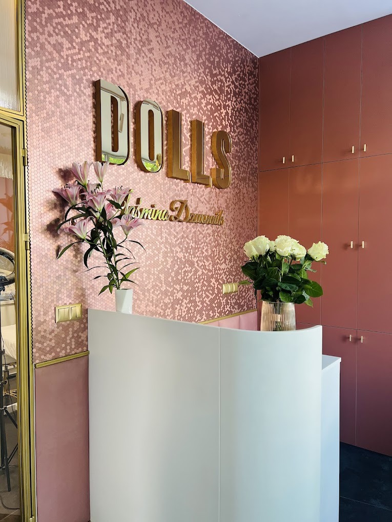 Eleganckie wnętrze: różowa mozaika heksagonalna na ścianie, złote litery 'DOLLS', biała lada recepcyjna z kwiatami, szafa w odcieniach różu.