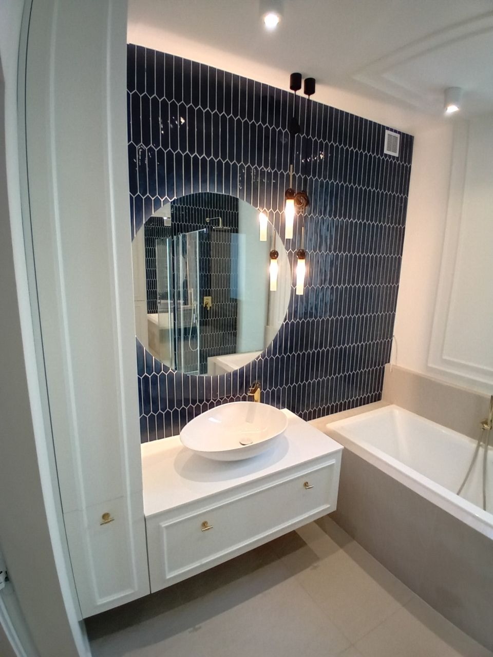 Elegancka łazienka po remoncie: ciemnoniebieska ściana z mozaiką, okrągłe lustro, biała umywalka nablatowa i wanna. Nowoczesne oświetlenie.
