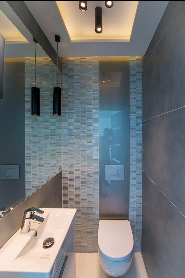 Nowoczesna łazienka z mozaiką w odcieniach błękitu i szarości, podwieszana toaleta, umywalka z baterią, oświetlenie punktowe i lustro. Minimalistyczny design.