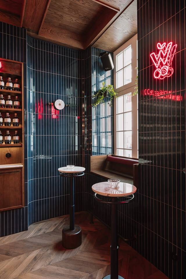 Stylowe wnętrze baru z ciemnymi, pionowo ułożonymi płytkami, drewnianą podłogą w jodełkę i neonowym napisem. Widoczne stoliki barowe i okno z rośliną.