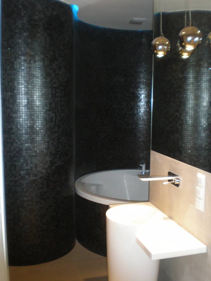 Nowoczesna łazienka z czarną mozaiką na zaokrąglonych ścianach, białą umywalką na postumencie i designerską armaturą. Minimalistyczny styl, oświetlenie punktowe.