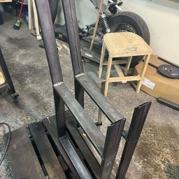 Podpergolą Mikołaj Szybka - Metalowa konstrukcja krzesła w trakcie spawania, widoczne spawy, surowy metal, warsztatowe otoczenie, paleta jako podstawa, w tle motocykl.