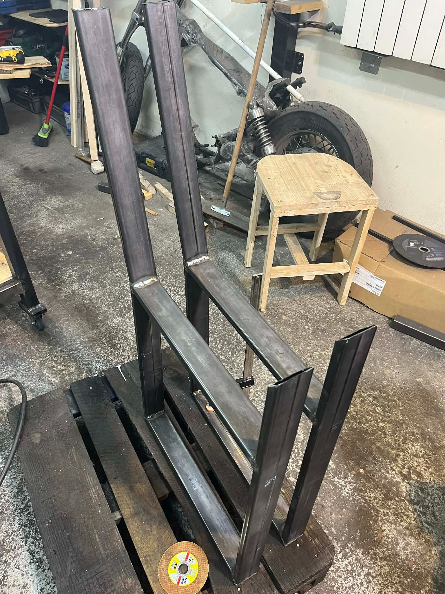 Metalowa konstrukcja krzesła w trakcie spawania, widoczne spawy, surowy metal, warsztatowe otoczenie, paleta jako podstawa, w tle motocykl.