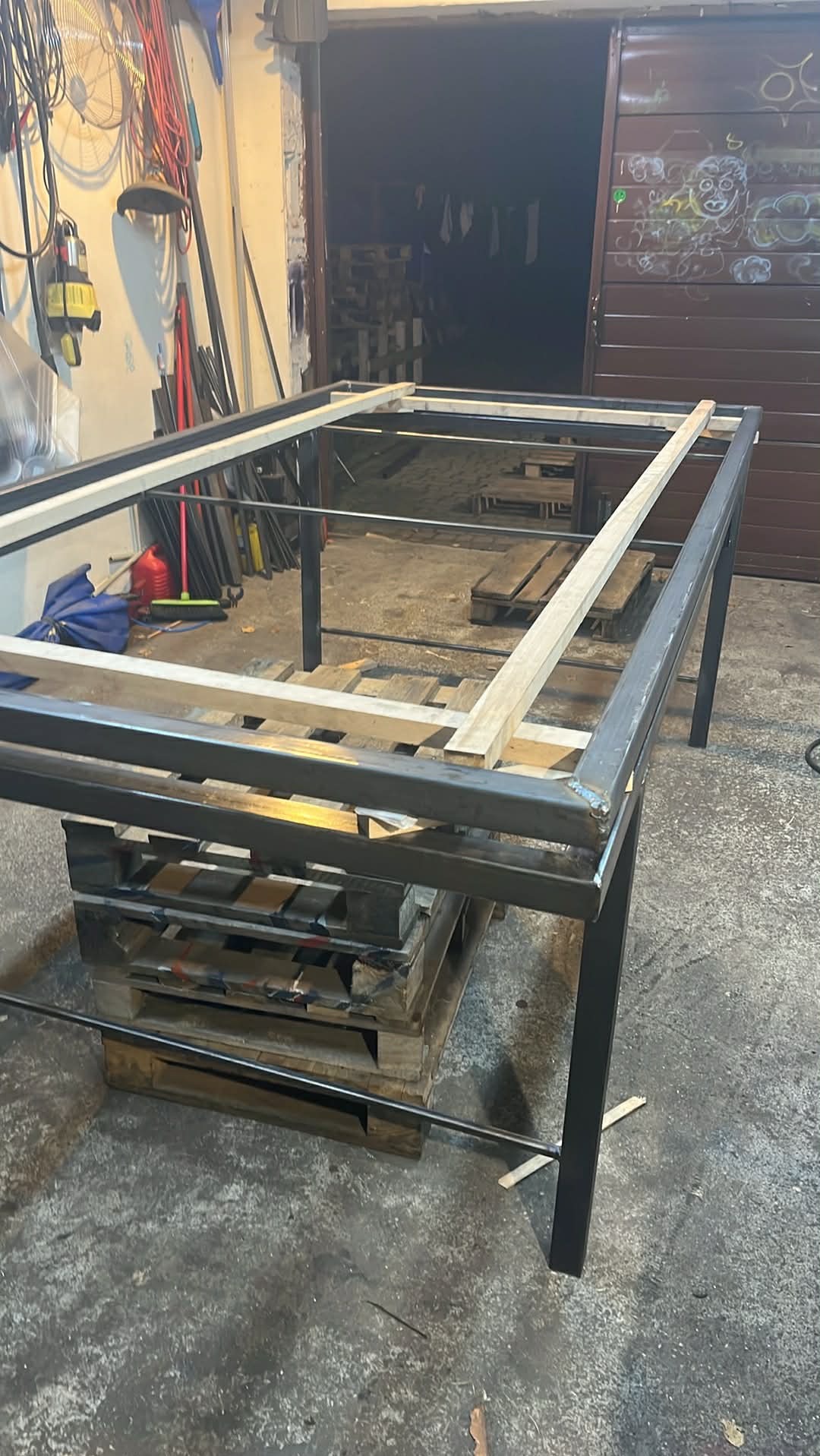 Stelaż metalowy z drewnianymi elementami, spawany, ustawiony na paletach w warsztacie. Widoczne ślady spawania i surowy wygląd konstrukcji.