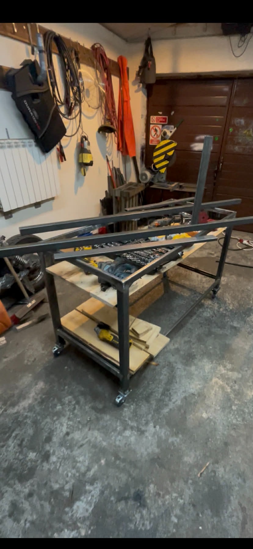 Stół spawalniczy w trakcie budowy, z narzędziami na półkach, w warsztacie. Widoczne elementy konstrukcyjne i narzędzia.