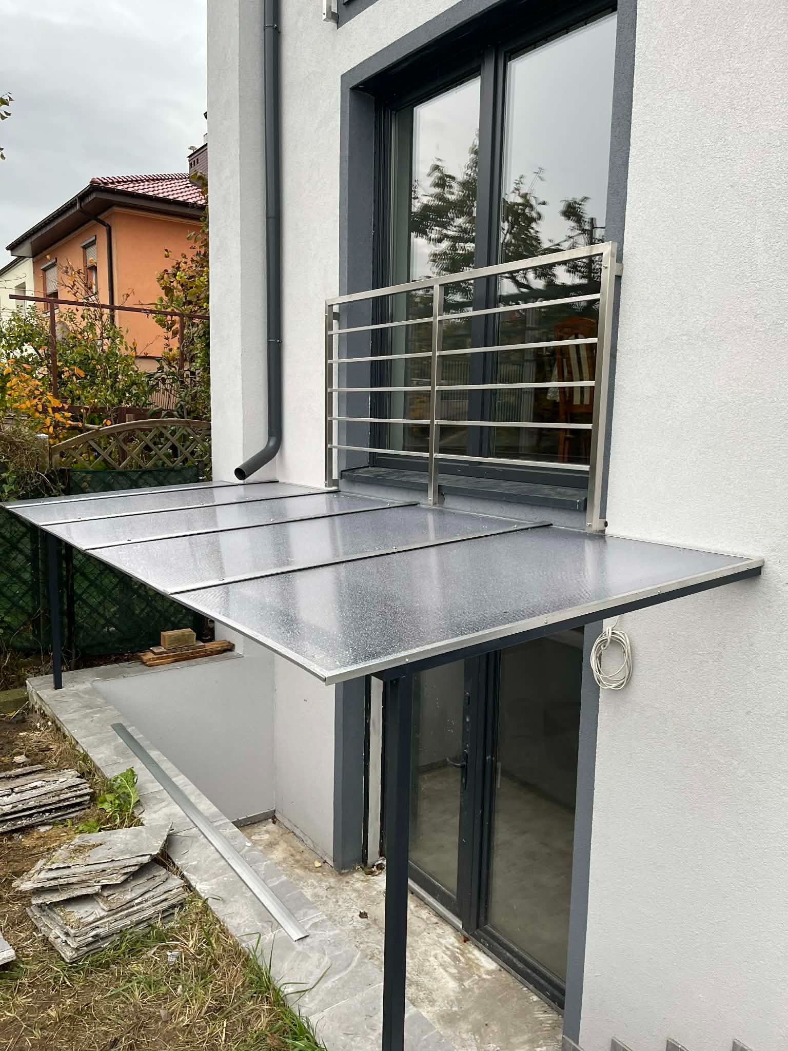 Zadaszenie nad zejściem do piwnicy z balustradą w Ostrzeszowie. Konstrukcja metalowa z przezroczystym pokryciem, widoczne detale wykończenia i otoczenie budynku.