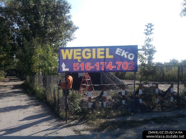 Tablica reklamowa z napisem 'Węgiel Eko' i numerem telefonu, widoczna z ulicy, za nią pryzma węgla i drabina.