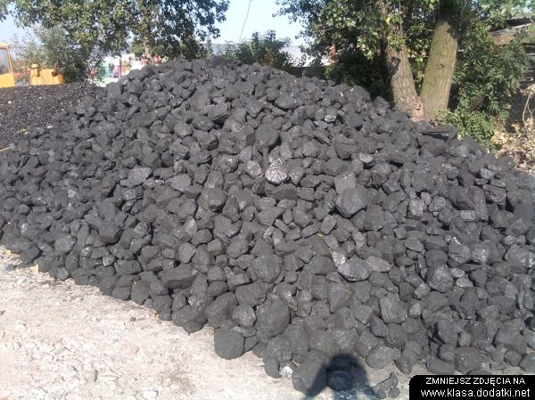 Kopa czarnego węgla kamiennego typu orzech II na placu składowym, z żółtą maszyną w tle i drzewami w tle.