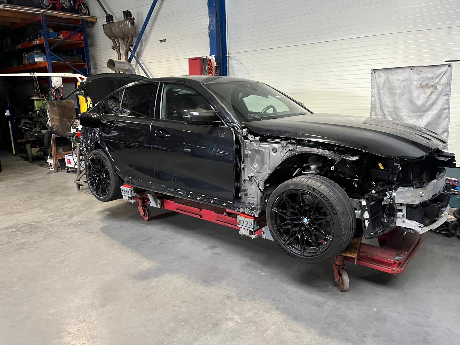 Czarny samochód na czerwonym podnośniku warsztatowym, zdemontowane elementy karoserii, widoczne ślady napraw blacharskich. Warsztat z wyposażeniem w tle.